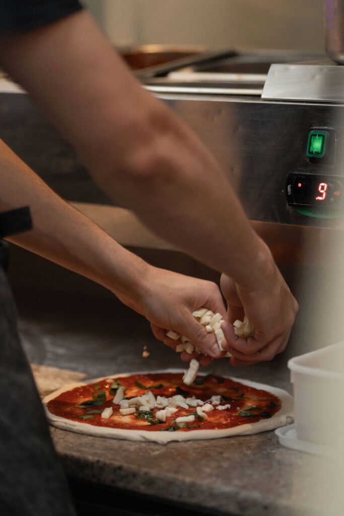 Close-up of Full Circle Brew Co pizza chefs sprinkling cheese on an uncooked pizza, preparing a delicious wood-fired creation for a wedding celebration.